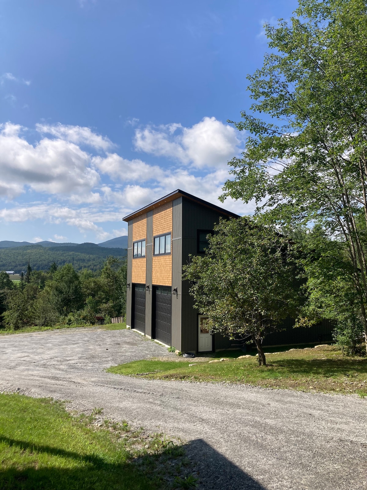 A two-story apartment building is set against a backdrop of rolling hills and blue skies. The structure features a mix of wood and metal siding, with large windows allowing natural light to fill the interiors. Surrounding greenery adds to the tranquil atmosphere.
