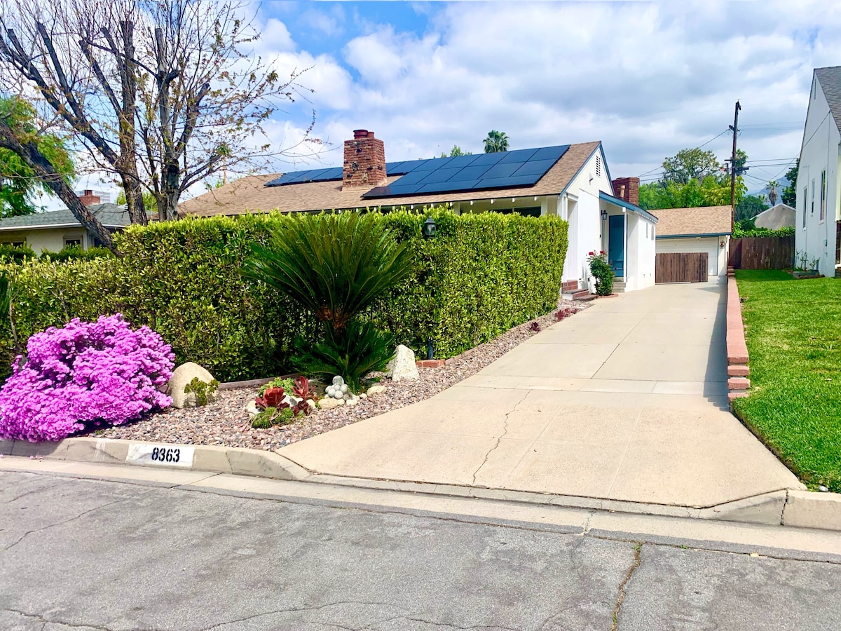 A well-maintained exterior of a residential property is shown, featuring a neat driveway leading up to the house. Lush greenery and decorative plants, including prominent purple flowers, frame the space. Solar panels are visible on the roof, and a clear sky complements the scene.