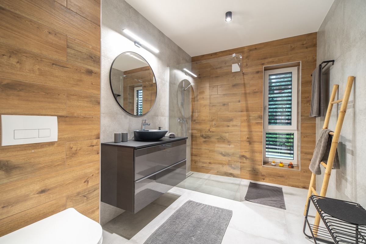 The bathroom features a modern design with wooden wall accents and sleek fixtures. A circular mirror is positioned above a dark vanity, complemented by a matching black sink. Natural light enters through a window adorned with blinds, highlighting the walk-in shower and neatly arranged towels.