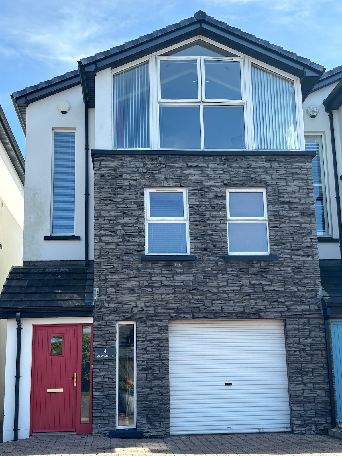 The exterior of the townhouse showcases a modern design with a stone-finished lower level and large windows above, allowing ample natural light. A vibrant red door provides a welcoming entry, complemented by a garage door. The neatly paved area in front features a clean, tidy appearance.