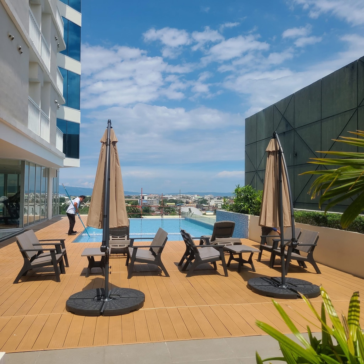 An outdoor area features a wooden deck with several comfortable lounge chairs and two large umbrellas. A swimming pool is visible in the background, accompanied by lush greenery and a clear sky, providing a serene atmosphere for relaxation.