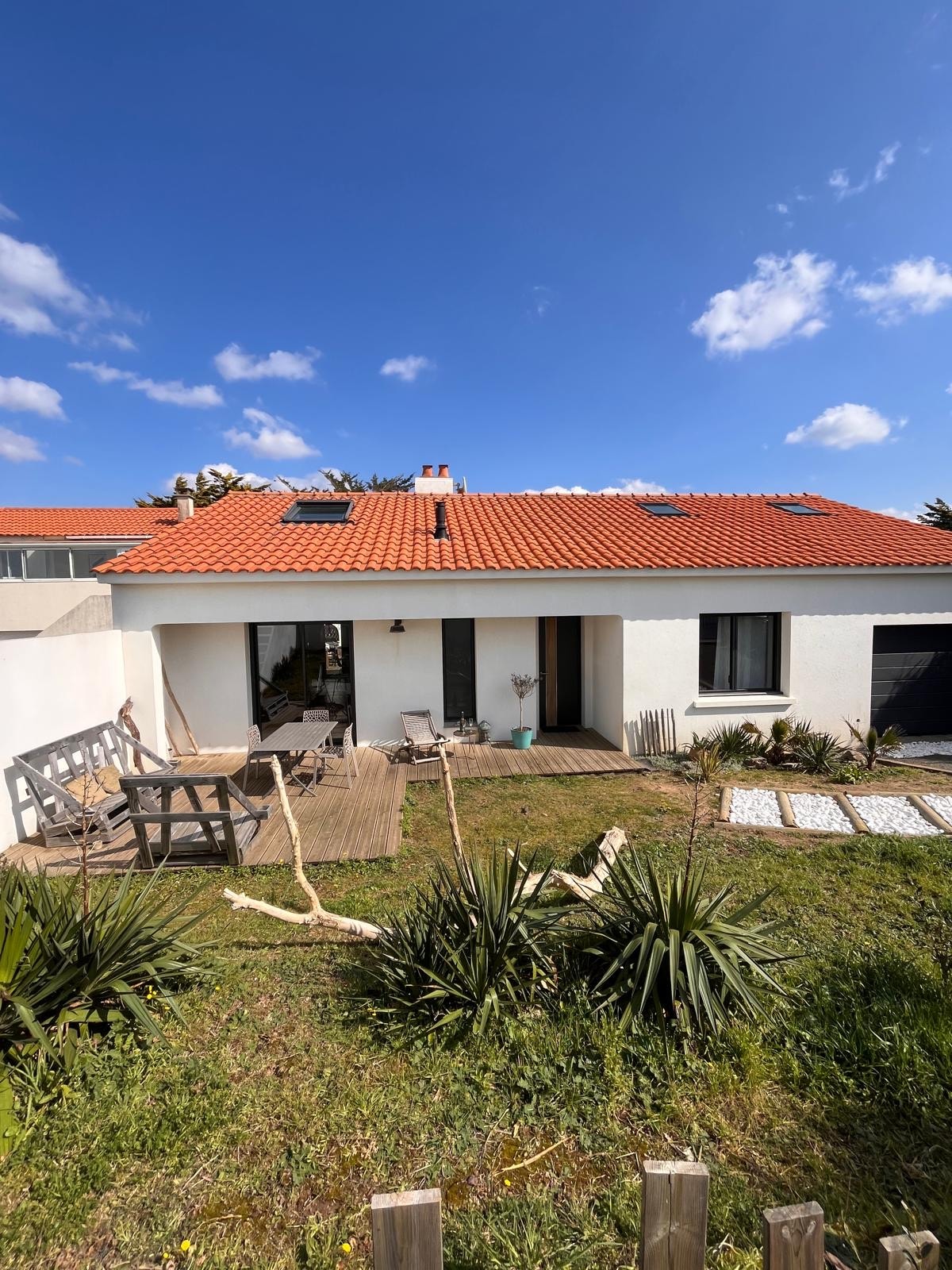 A modern villa exterior is displayed under a clear blue sky. The white facade features dark shutters and a welcoming entrance. A wooden deck is complemented by outdoor seating and landscaped greenery, creating a tranquil outdoor space. Details of the roof and nearby plants are also visible.