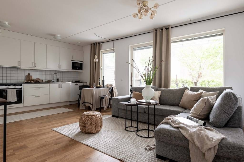 A living area features a spacious gray sofa complemented by a cozy knit throw. A coffee table, accompanied by two smaller tables, sits on a woven rug. Natural light enters through large windows framed by light curtains, illuminating the open kitchen with modern cabinetry.