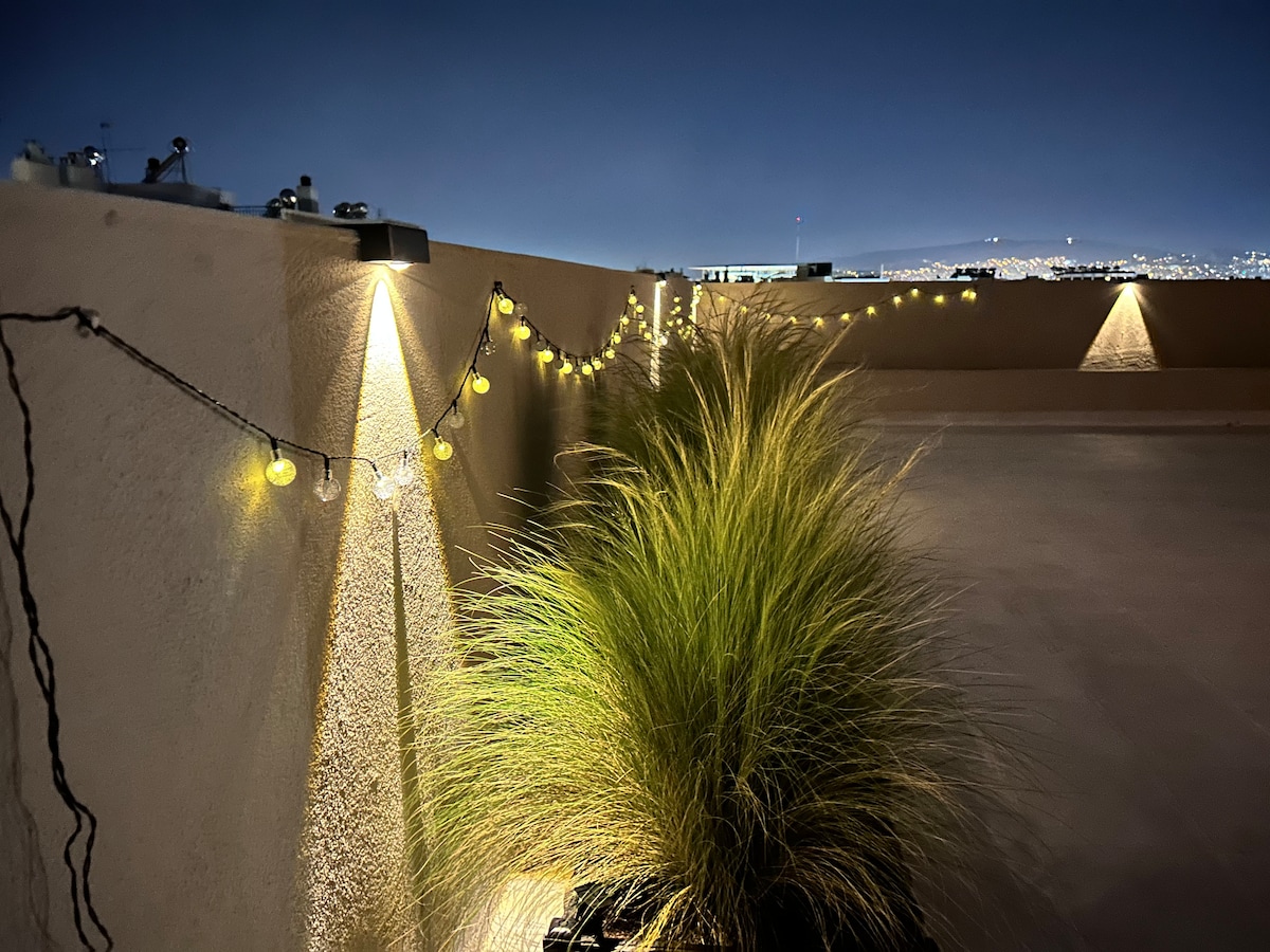 A balcony is illuminated by soft yellow string lights, creating a relaxing ambiance. Lush green grasses are also present, contributing to the serene environment. In the background, distant city lights can be seen, further enhancing the evening atmosphere.