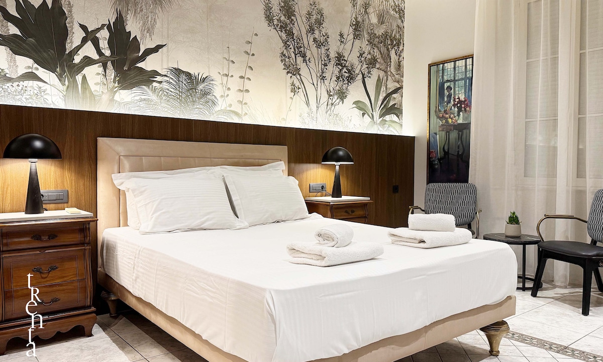 A spacious bedroom features a large bed with neatly arranged white linens and towels. Soft lighting is provided by two bedside lamps. Behind the bed, an artistic mural of plants adds a natural element to the decor, complemented by a small seating area with two chairs.