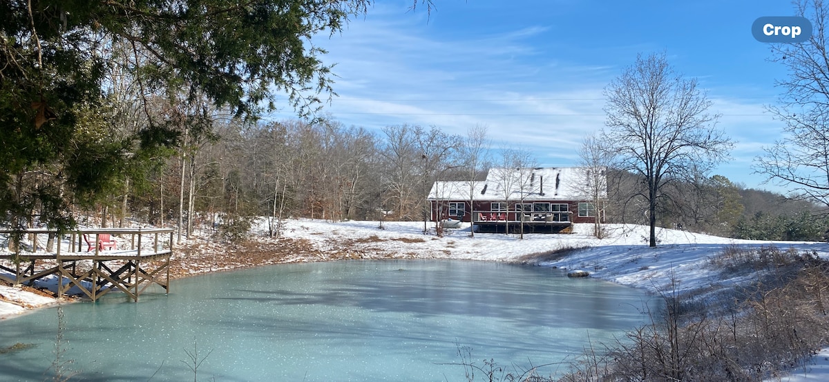 A serene pond reflects a clear blue sky, with a modern farmhouse visible in the background. Snow lightly covers the ground and the roof, while bare trees are scattered throughout the landscape, complementing the tranquil setting.