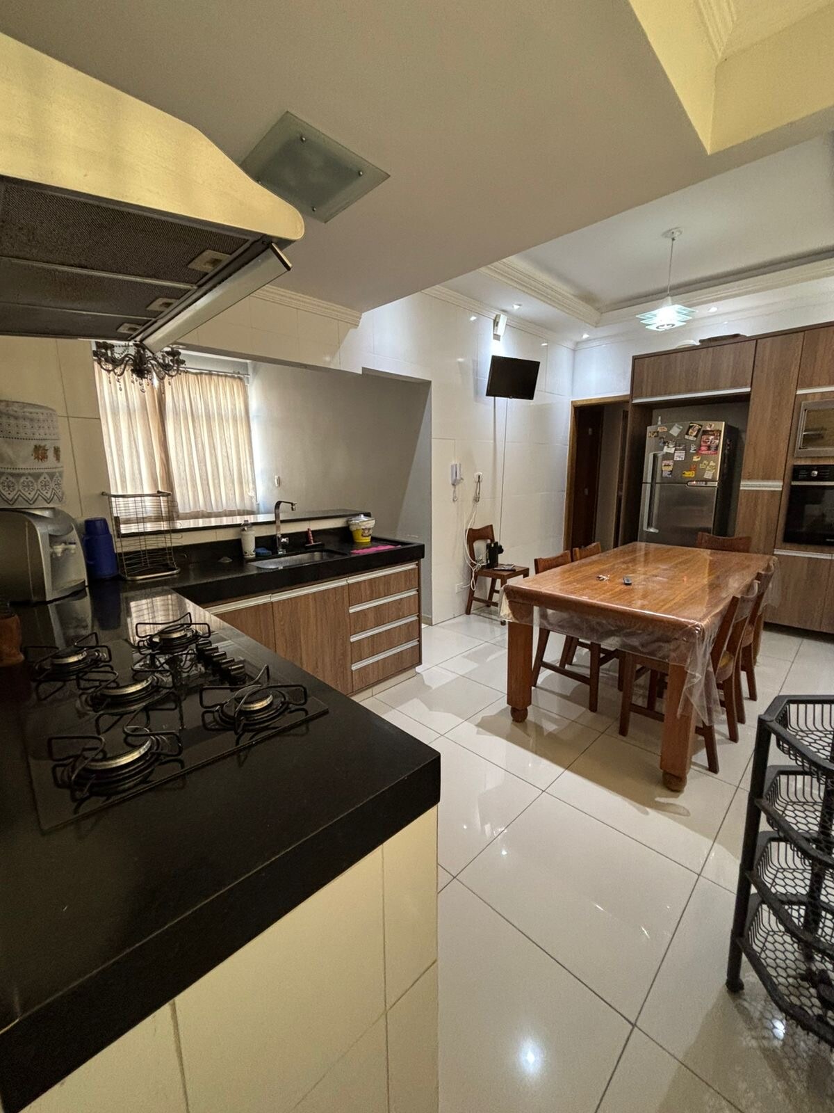 The kitchen features a modern design with dark countertops and light cabinetry. A large wooden dining table with eight chairs is visible, providing ample space for meals. Appliances include a stainless steel refrigerator and a stove with multiple burners. Natural light filters through a window covered by sheer curtains.