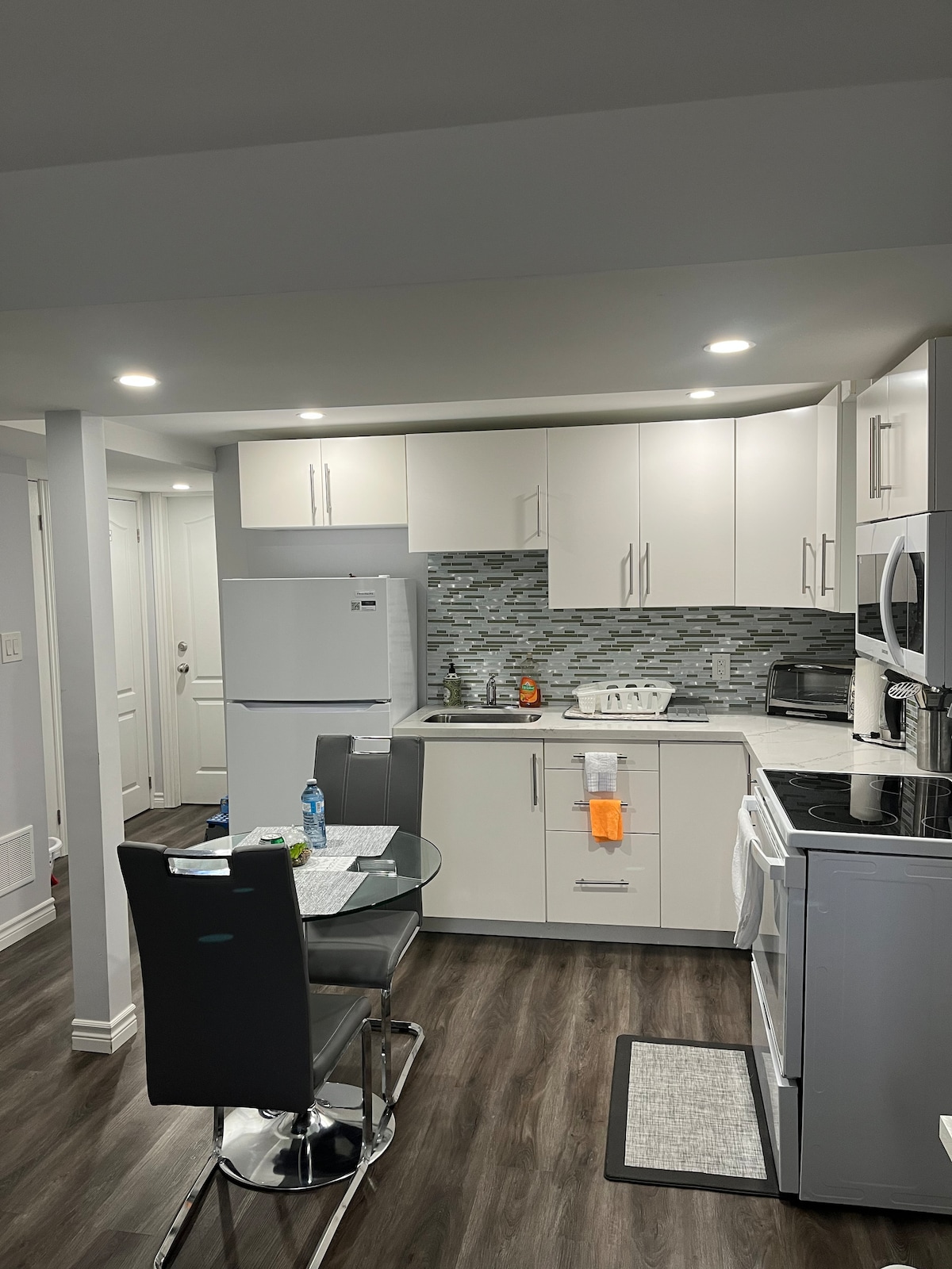 The kitchen area features modern cabinetry and a tiled backsplash. A round dining table with two black chairs is set beside a window. Stainless steel appliances, including a refrigerator and microwave, are visible, alongside a stove and additional counter space.