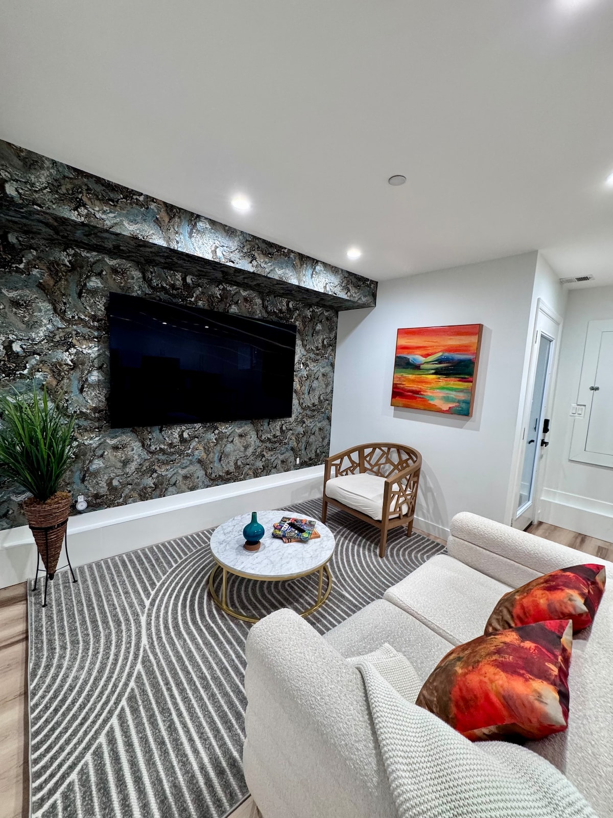 A modern living area showcases a large television mounted on a textured accent wall. A decorative circular table sits in front of a comfortable sofa adorned with colorful cushions. An area rug with a wavy pattern adds warmth, complemented by a potted plant and a colorful artwork.