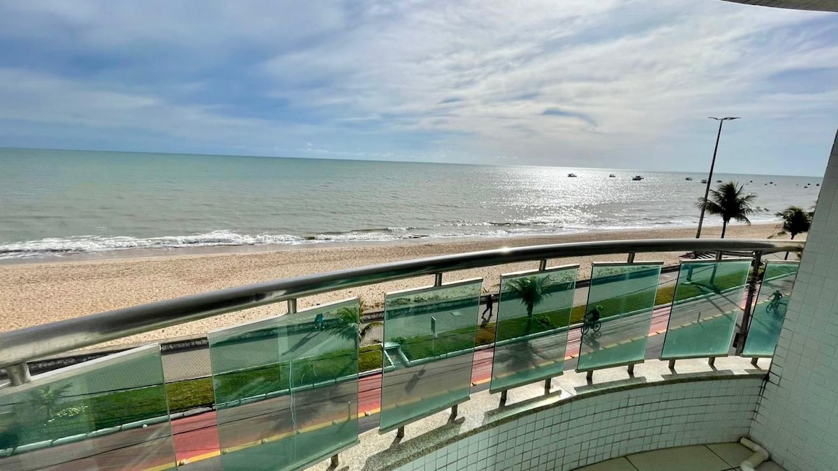 A spacious balcony presents a panoramic view of the ocean, featuring the beach and gentle waves in the distance. The railing is adorned with green-tinted glass panels, allowing for an unobstructed sight of the coastline and the horizon.