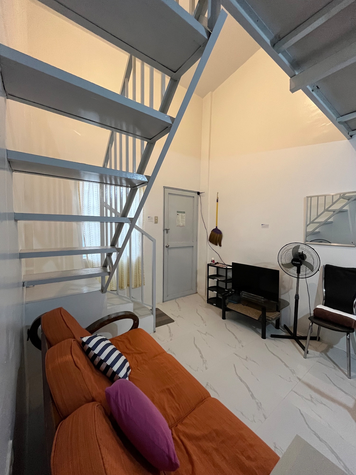 A loft-style living area features an orange sofa with a pink pillow, complemented by a simple side table. A small television unit is positioned nearby, with a floor fan for airflow. A staircase leads to the upper level, and natural light filters in through a window.