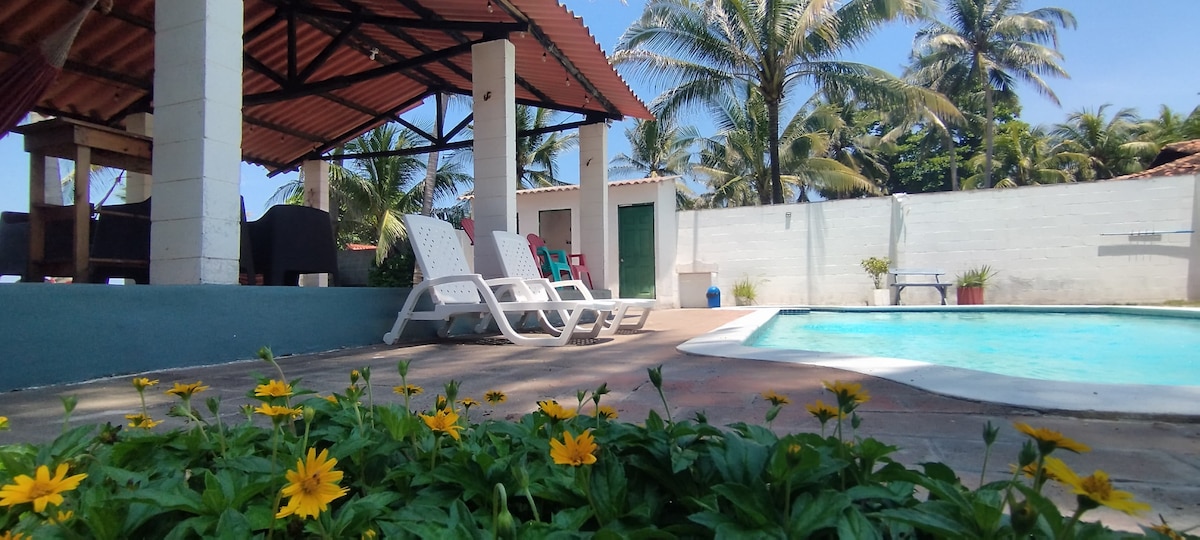 A refreshing pool is surrounded by a tiled area with lounge chairs positioned nearby. Bright yellow flowers bloom at the forefront, while a covered outdoor seating area and palm trees can be seen in the background under a clear blue sky.