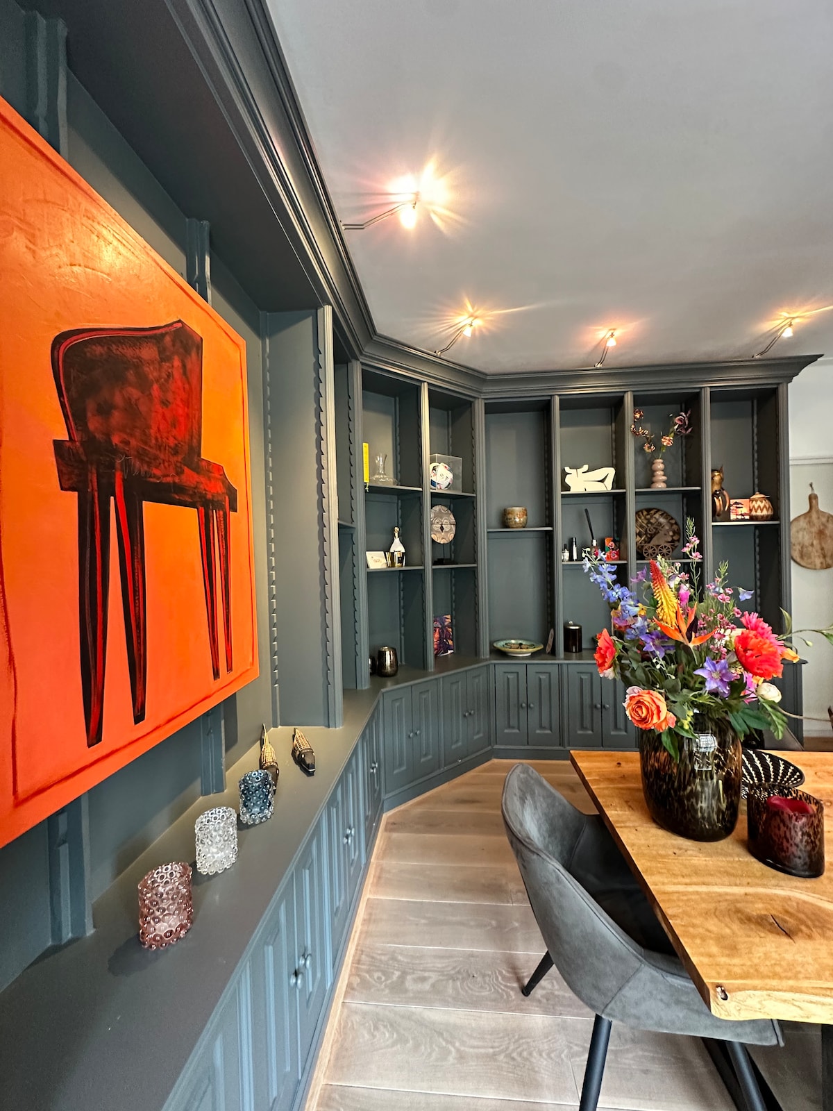 A modern dining area features a wooden table surrounded by gray chairs. A vibrant orange artwork of a chair is displayed on the wall, complemented by an array of decorative items on shelves. A colorful flower arrangement adds a touch of freshness to the space.