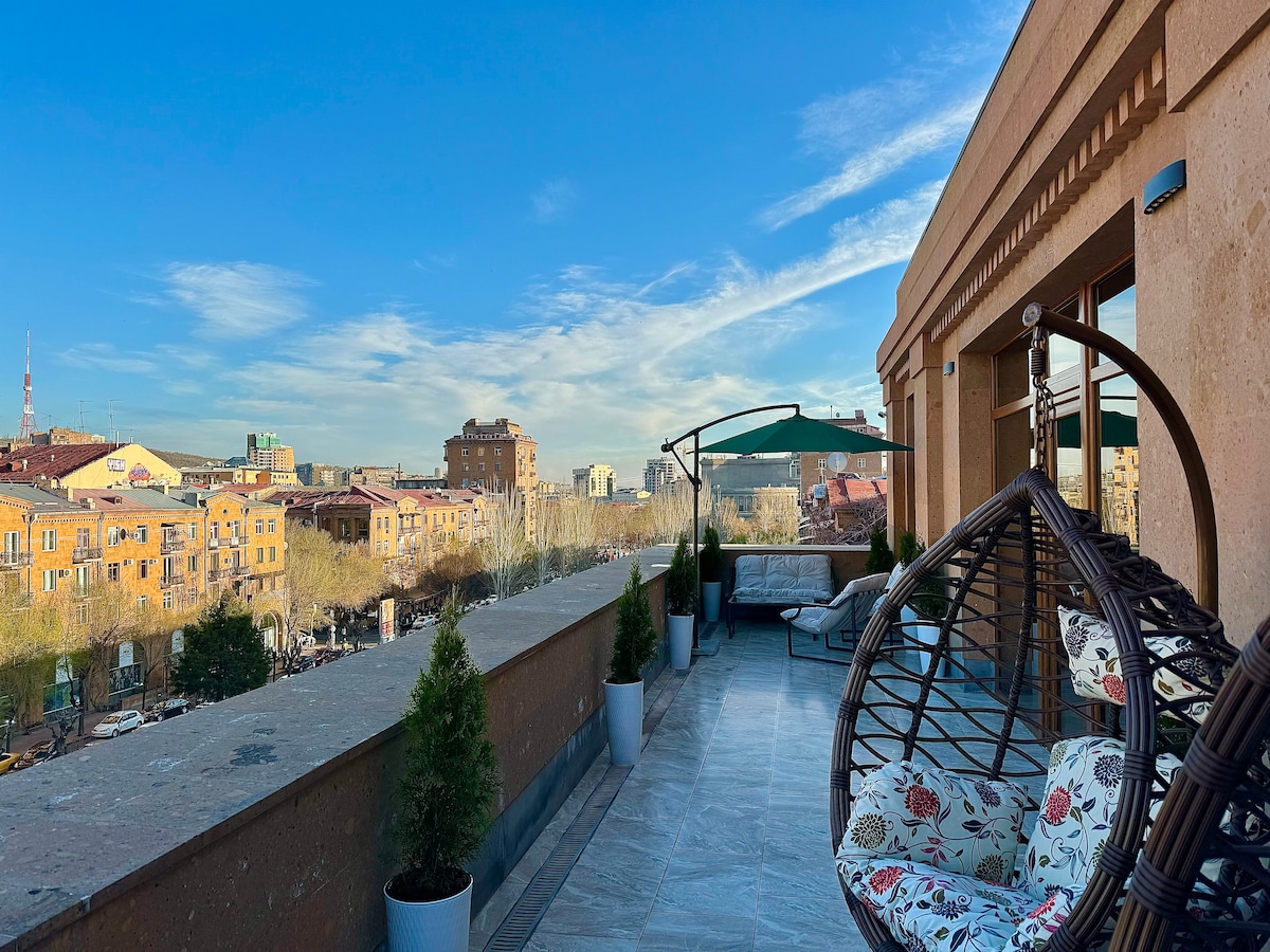 A spacious terrace features hanging chairs and lounge seating, surrounded by planters. A canopy provides shade while offering views of the cityscape and the sky. The design highlights a calm atmosphere, ideal for relaxing outdoors.