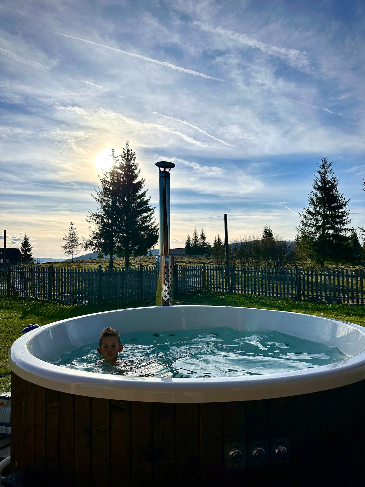 A round hot tub is situated outdoors, surrounded by greenery and a wooden fence. The sun sets in the background, casting warm light over the scene, while trees frame the view. The water in the hot tub reflects the sky's hues.