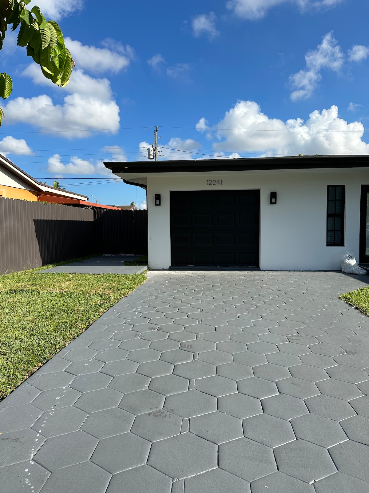 The exterior of the guesthouse is depicted, featuring a modern facade with a garage door and two narrow windows. A spacious driveway paved with hexagonal tiles leads to the entrance. Lush green grass borders the driveway, and a fence provides privacy.