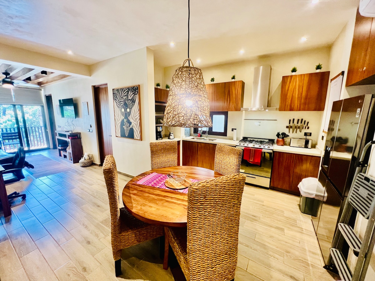 A modern kitchen area features wooden cabinetry and stainless steel appliances. Wicker chairs surround a round dining table, highlighted by a pendant light fixture above. A piece of artwork is displayed on the wall, and access to a balcony is visible in the background.