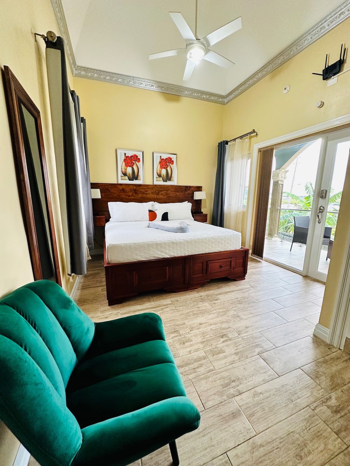 A spacious room features a queen bed with wooden furnishings and colorful artwork on the walls. A green accent chair is positioned nearby. Natural light filters in through glass doors leading to an outdoor area, with light-colored tiled flooring enhancing the bright atmosphere.