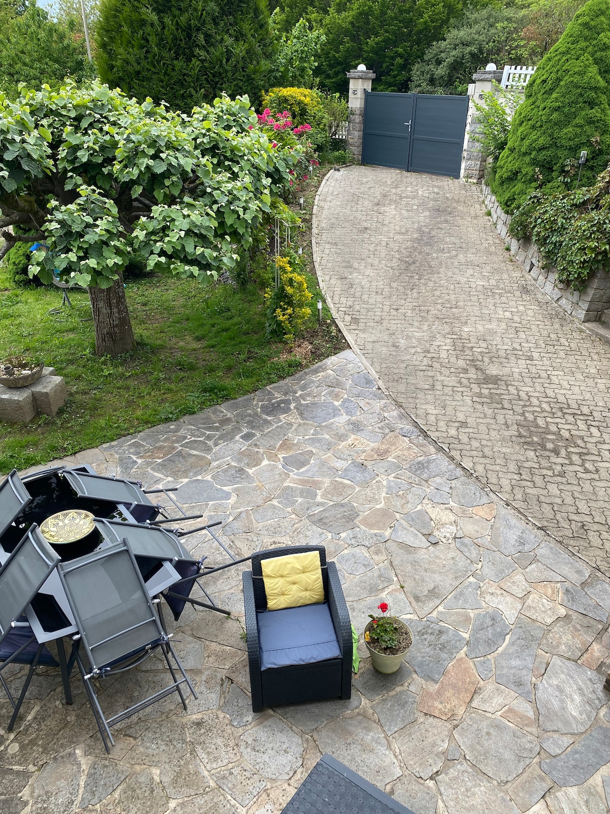 The outdoor area features a stone-paved terrace with a round table and several folding chairs. A single yellow cushion is placed on one chair. Lush greenery, including a tree and flowering plants, surrounds the space, while a private driveway leads to a gated entrance.