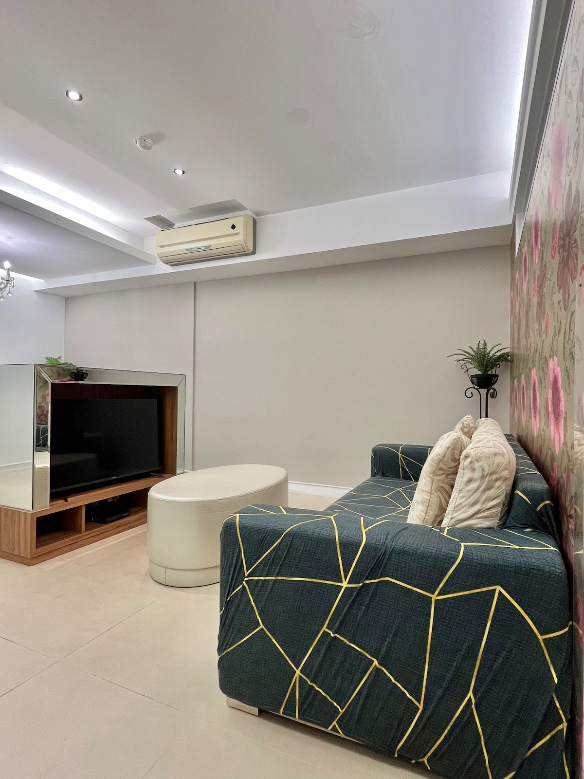 A modern living space is displayed, highlighting a cozy sofa with geometric patterns and decorative pillows. A sleek white coffee table sits in front of a stylish entertainment unit that houses a large flat-screen TV. Soft lighting and floral wallpaper add a touch of elegance.