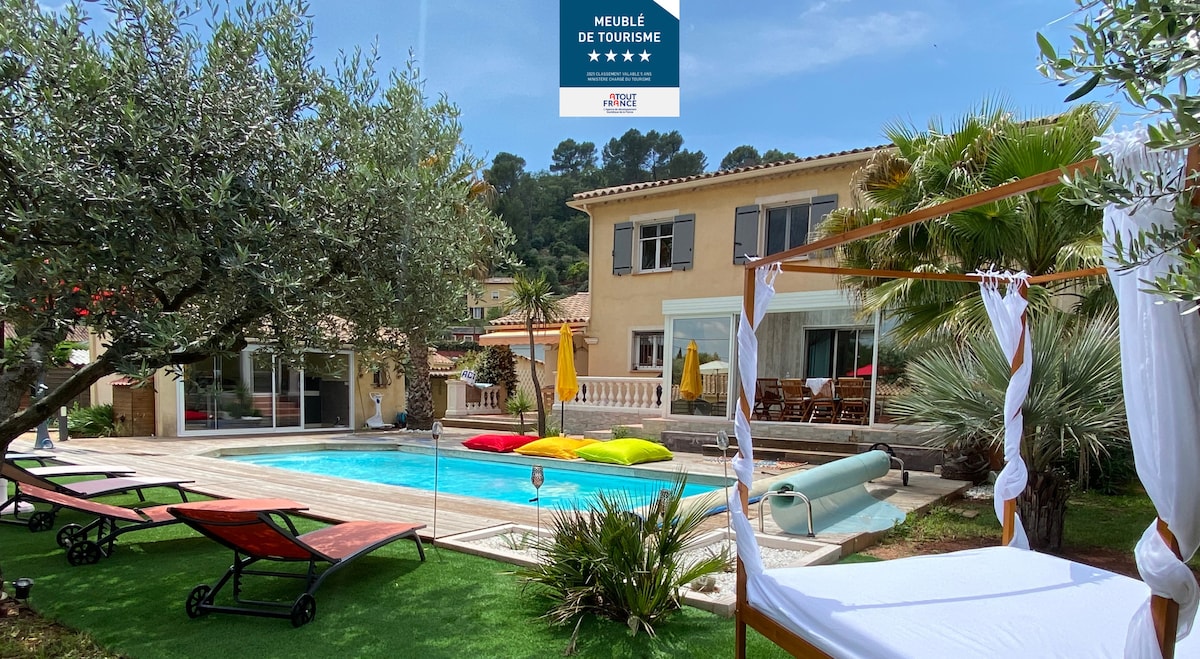 An inviting villa exterior showcases a heated pool surrounded by lush greenery and colorful lounging areas. Comfortable deck chairs and vibrant cushions are positioned near the poolside. The multi-story villa features a charming façade, with outdoor dining space visible on the terrace.