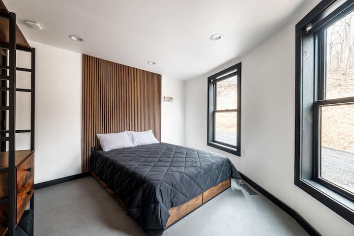 A bright bedroom with a wood-slat accent wall, platform bed, and two windows overlooking the woods.