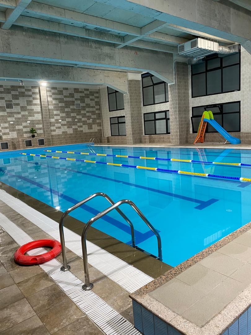 A spacious indoor swimming pool is framed by high, bright windows, allowing natural light to fill the area. Colorful lane markers are arranged across the water, with a yellow slide positioned nearby. A lifebuoy rests on the tiled deck next to steps leading into the pool.