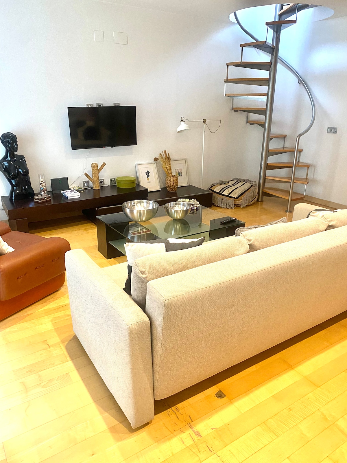 A stylish living area features a spacious sofa facing a wall-mounted television. A coffee table with decorative items is centered in the room, while a modern spiral staircase leads to an upper level. Warm wood flooring complements the neutral palette.