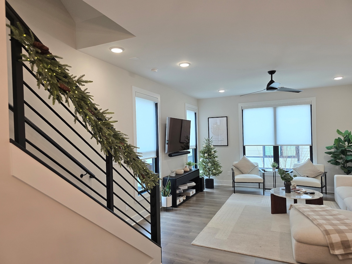 The living area is showcased with a light-filled layout featuring a spacious couch, accent chairs, and a round coffee table. A mounted TV and decorative plants add to the modern ambiance, while a tasteful garland adorns the staircase railing.