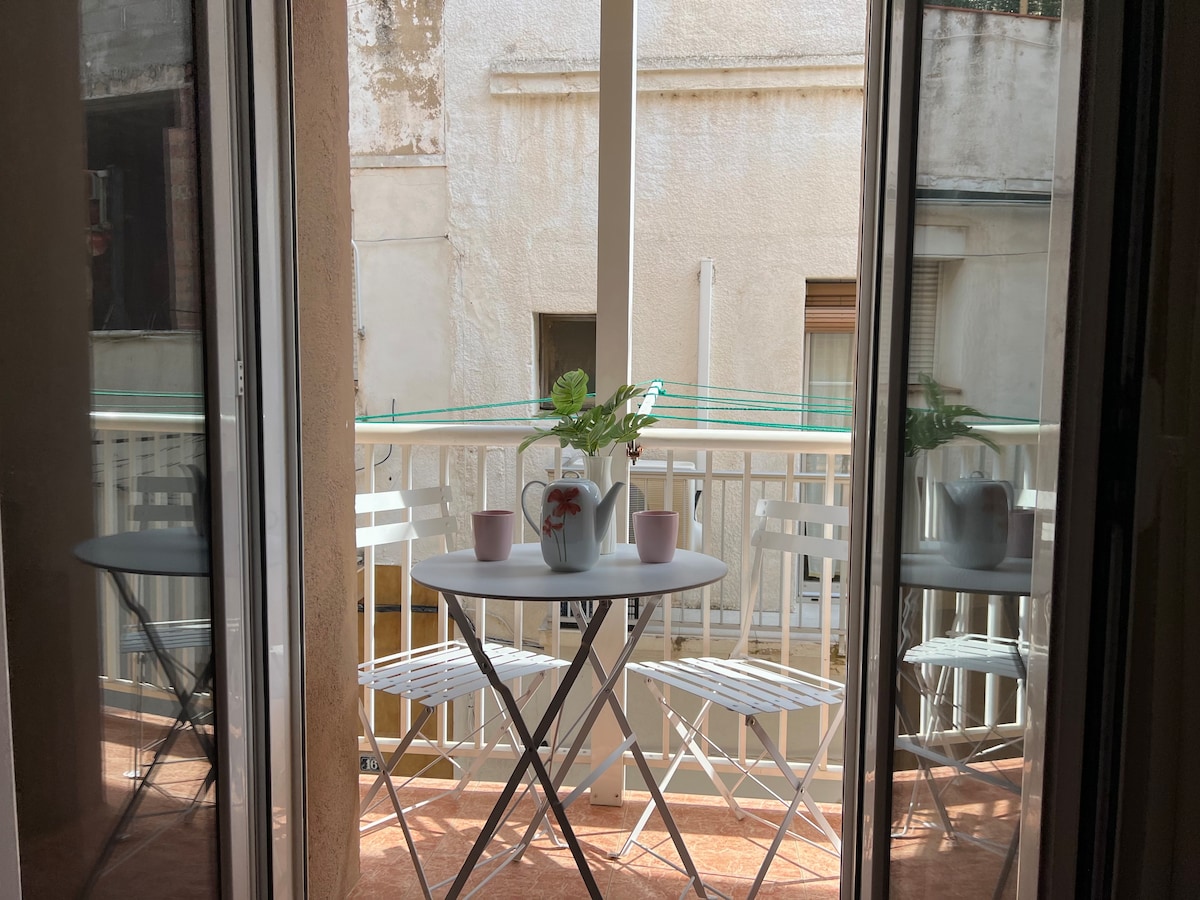 A small outdoor balcony area is depicted, featuring a round table set for two with white folding chairs. A potted plant and two pink cups are positioned on the table, creating a serene spot for relaxation. Glass doors frame the view, allowing natural light to enter.
