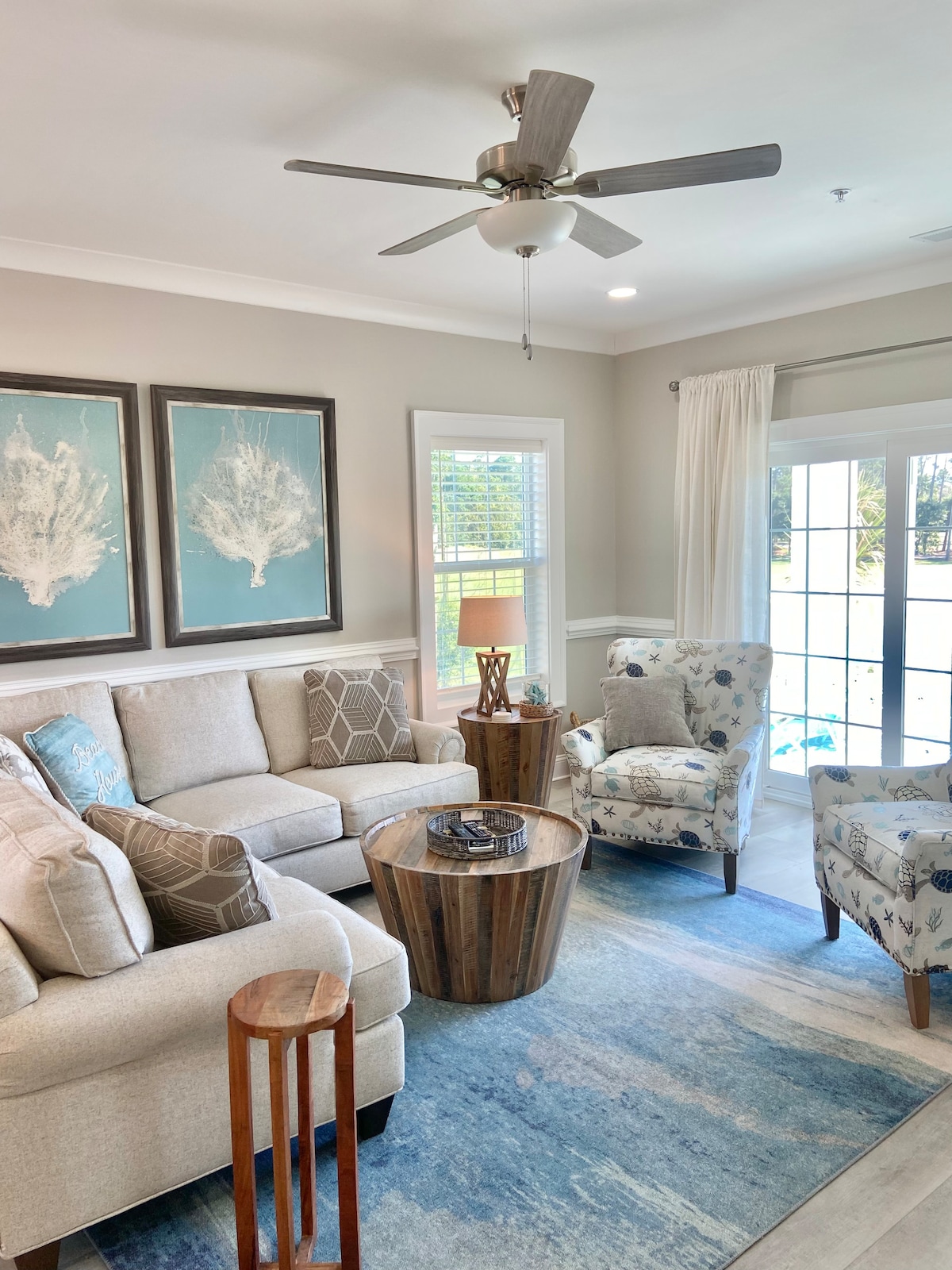 A comfortable living area features a light-colored sofa and two stylish accent chairs. A wooden coffee table sits at the center, accompanied by a small side table. Natural light enters through a set of glass doors, framed by sheer curtains, enhancing the inviting ambiance.