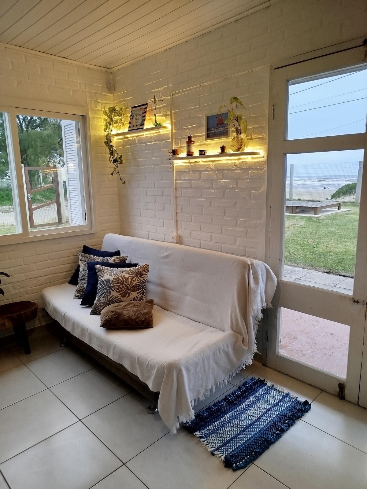 A cozy seating area features a white-covered couch adorned with various decorative pillows. Soft lighting is provided by illuminated shelves holding small decor items. A large window allows natural light, with a view of the grassy outdoor space and the sea visible beyond.