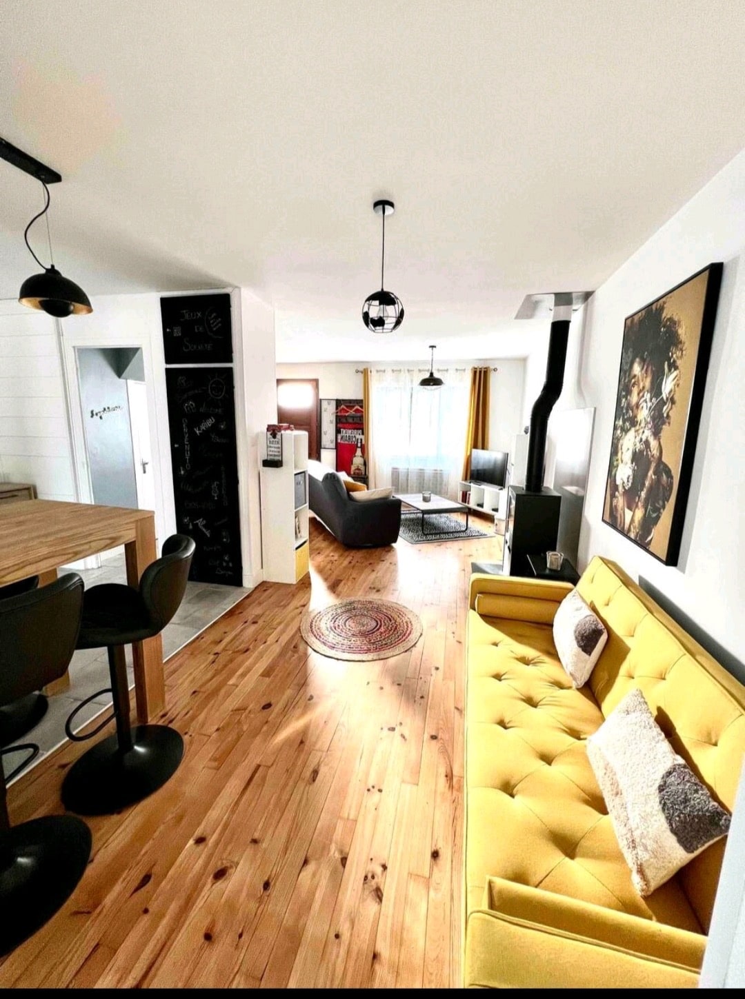 An open living space showcases a bright yellow sofa paired with dark armchairs. A wooden dining table with high stools is present, along with a round rug on the polished wooden floor. Large windows allow natural light to fill the area, enhancing the overall atmosphere.