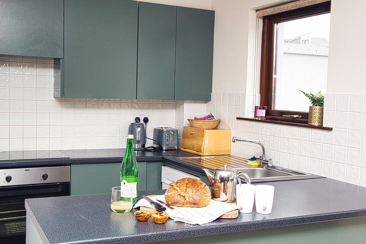 A functional kitchen is visible, featuring dark countertops and modern green cabinetry. A loaf of bread is positioned on a cloth next to glassware and a bottle of sparkling water. Kitchen appliances, including a toaster and kettle, are neatly arranged, with natural light coming from a nearby window.