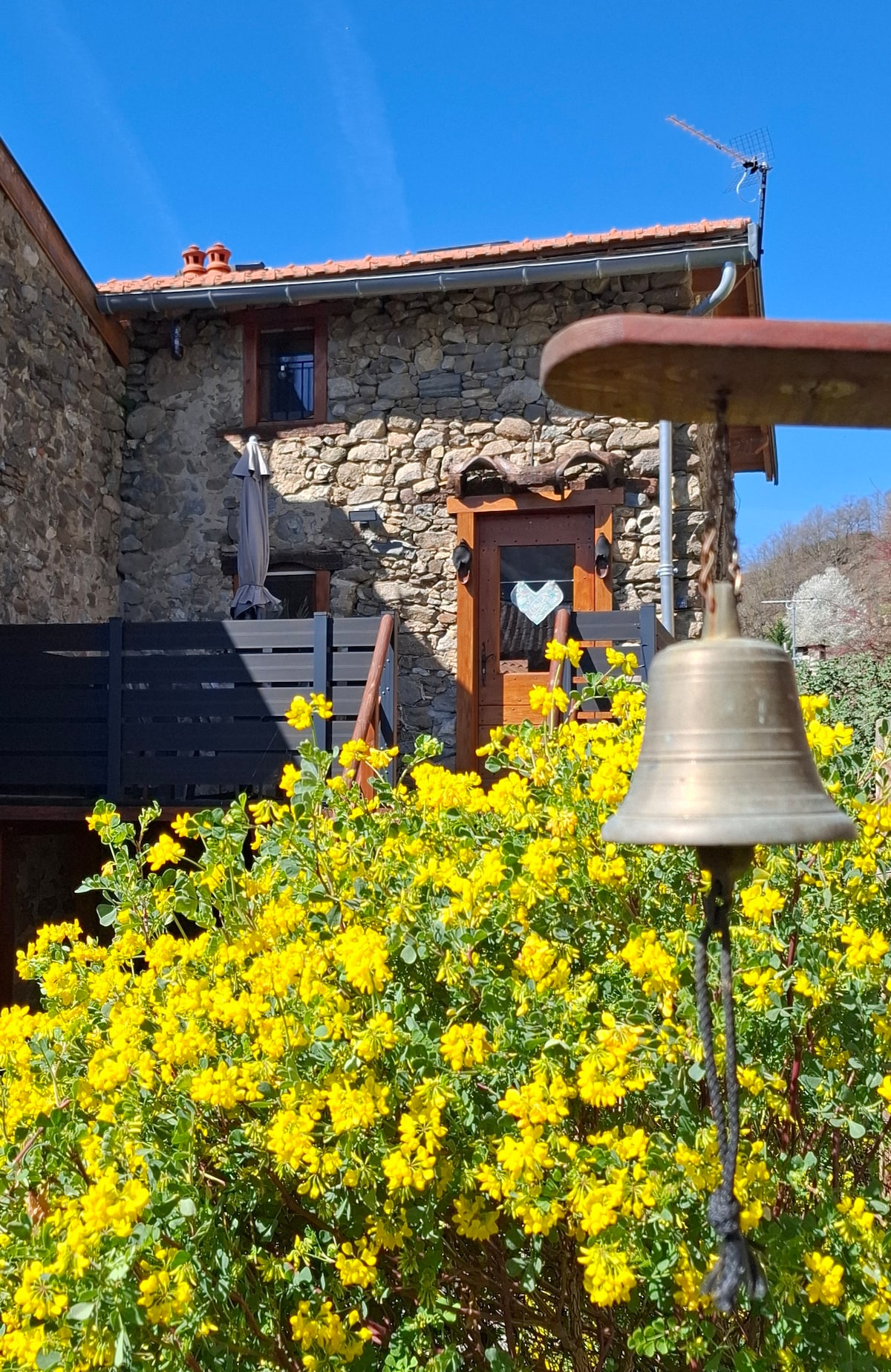 A stone house stands with a wooden door adorned by a heart shape, framed by a vibrant yellow flower bush. The balcony above features smooth wooden railings, and a bell hangs prominently in the foreground, adding a rustic charm to the scene.
