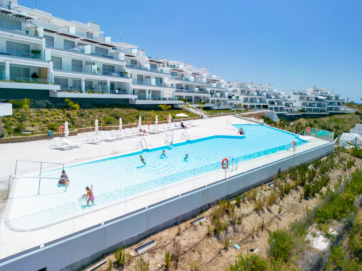 A large outdoor swimming pool is featured, surrounded by spacious sunbathing areas and lounge chairs. Several guests are enjoying the water, with modern apartment buildings visibly positioned in the background. Lush landscaping borders the pool area, contributing to a serene environment.