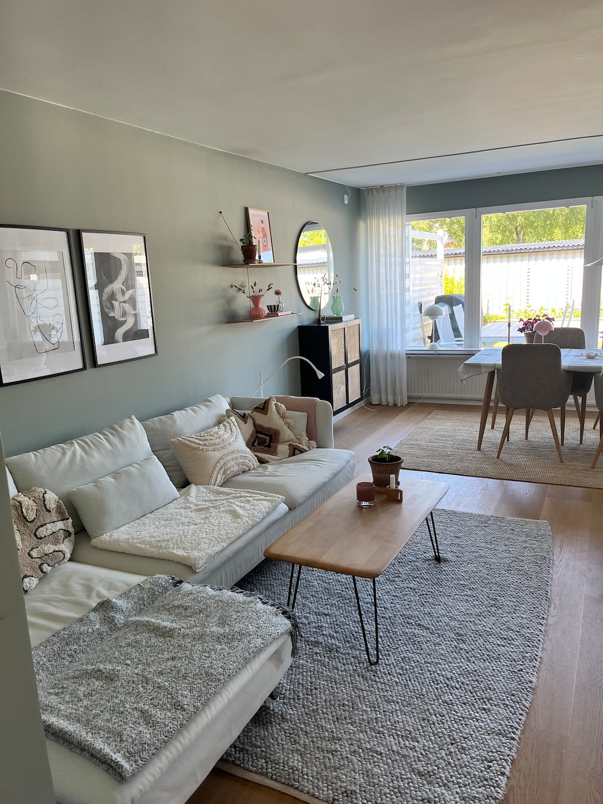An inviting living area features a light-colored sofa adorned with decorative pillows and throws. A wooden coffee table is placed in front, while shelves display various plants and artwork. Large windows allow natural light to fill the space, with a dining area visible in the background.