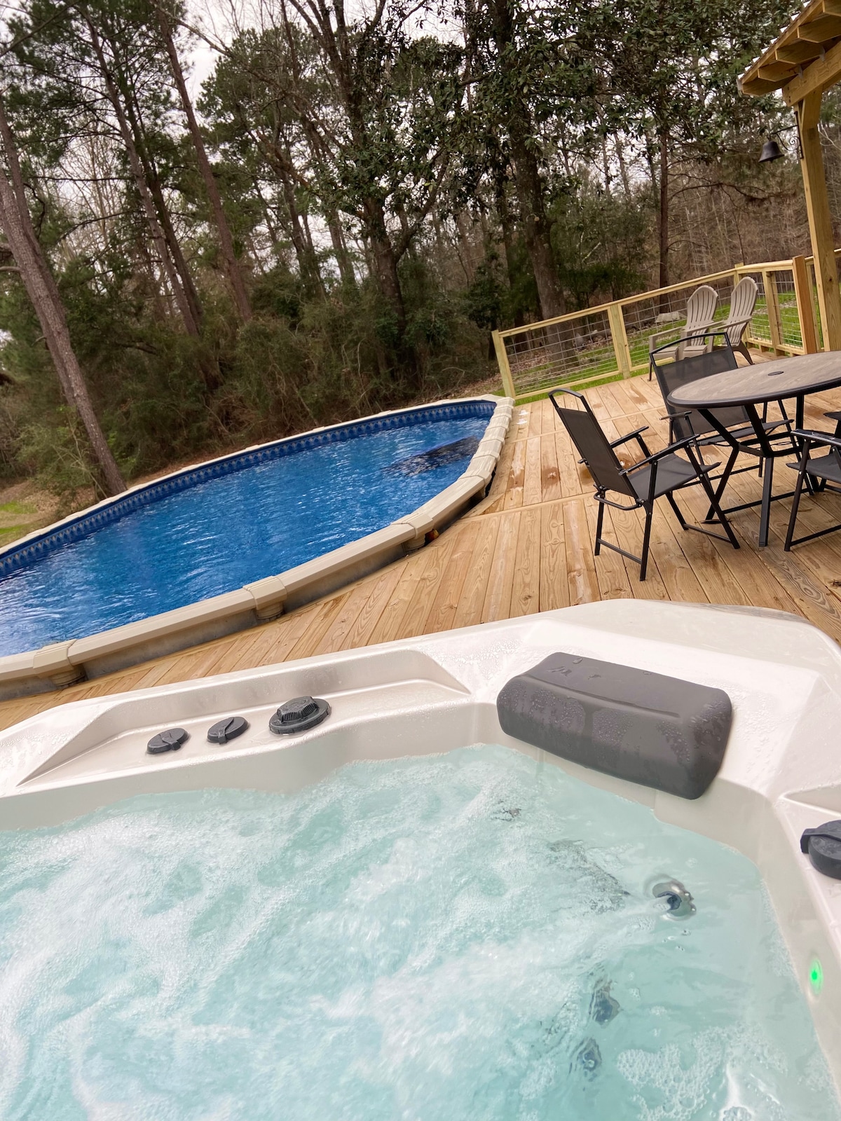 A hot tub is positioned on a wooden deck, with bubbling water inviting relaxation. Nearby, a swimming pool with a clear, blue surface reflects the surrounding trees. Outdoor seating is arranged around a table, providing space for gatherings.