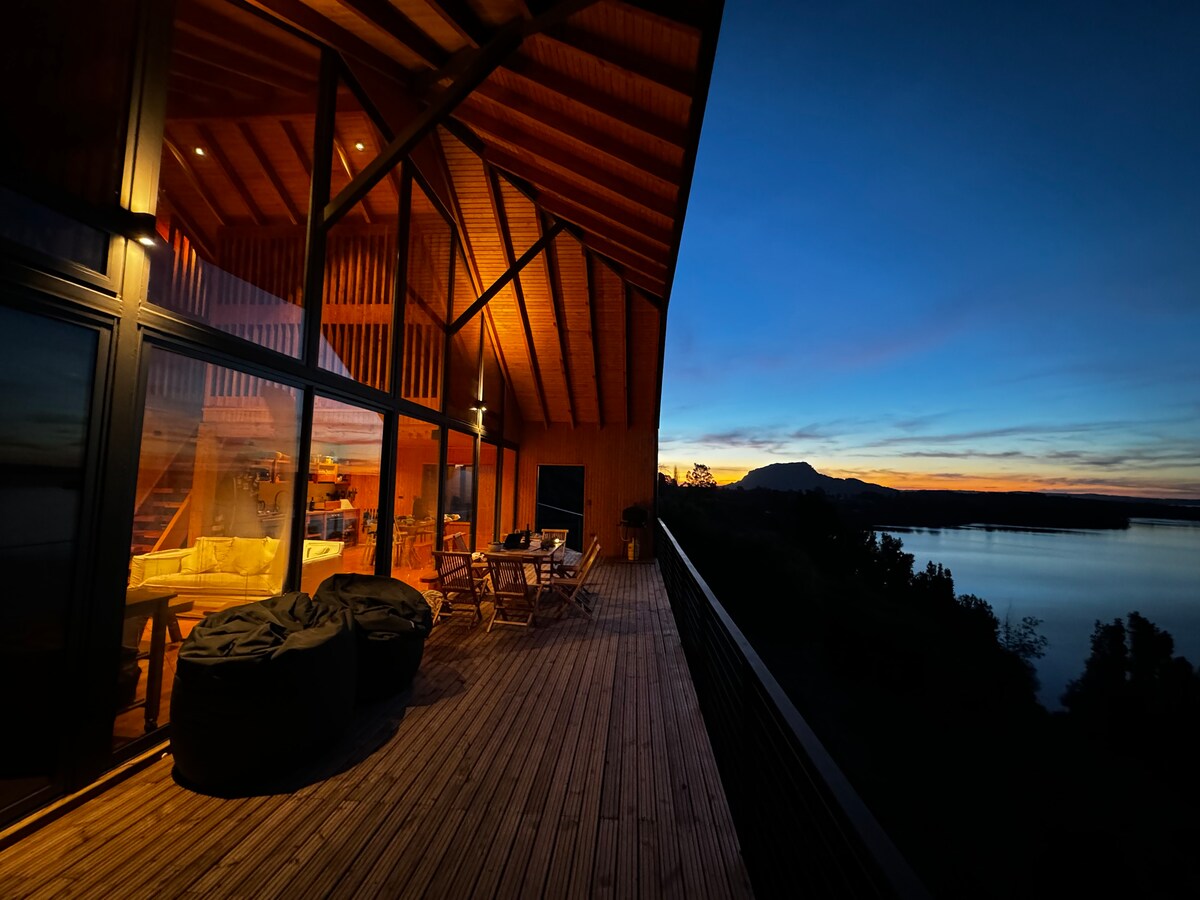 An expansive wooden deck showcases a view of the lake at dusk, with warm lighting highlighting the interior space. A dining table and seating area are visible, framed by large windows that connect the living space to the outdoors. The silhouette of mountains is seen in the distance.