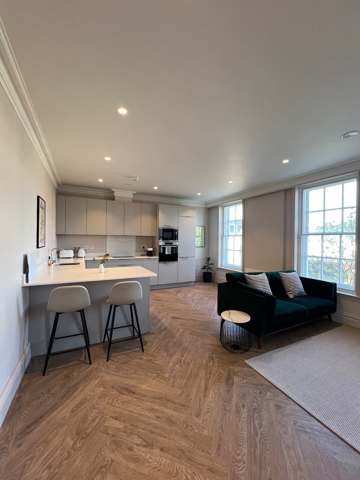 An open plan kitchen and living area is featured, showcasing a modern design with light cabinetry and a breakfast bar with two stools. A comfortable sofa is positioned near large windows, allowing natural light to illuminate the space. Wooden flooring adds warmth to the room.