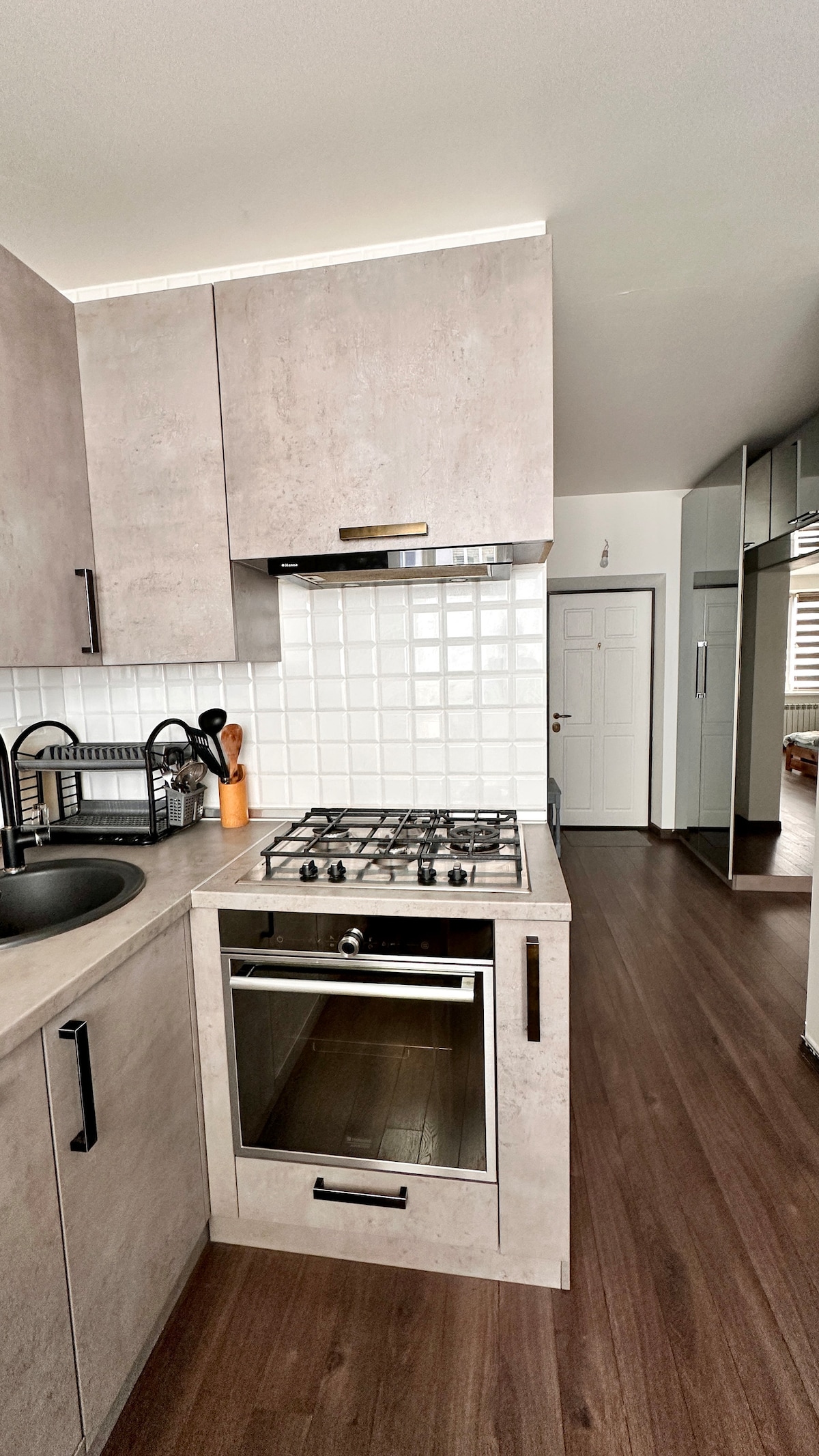 A modern kitchen corner features gray cabinetry and a white tile backsplash. A gas stove and oven are positioned under a functional range hood. Kitchen utensils are neatly organized, while the flooring displays a warm wood finish. A doorway leads to another room, visible in the background.