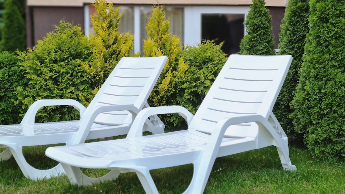 Two white lounge chairs are positioned on a green lawn, surrounded by neatly trimmed shrubs. The bright chairs offer a comfortable seating option, with the backdrop featuring large windows that allow natural light into the space.