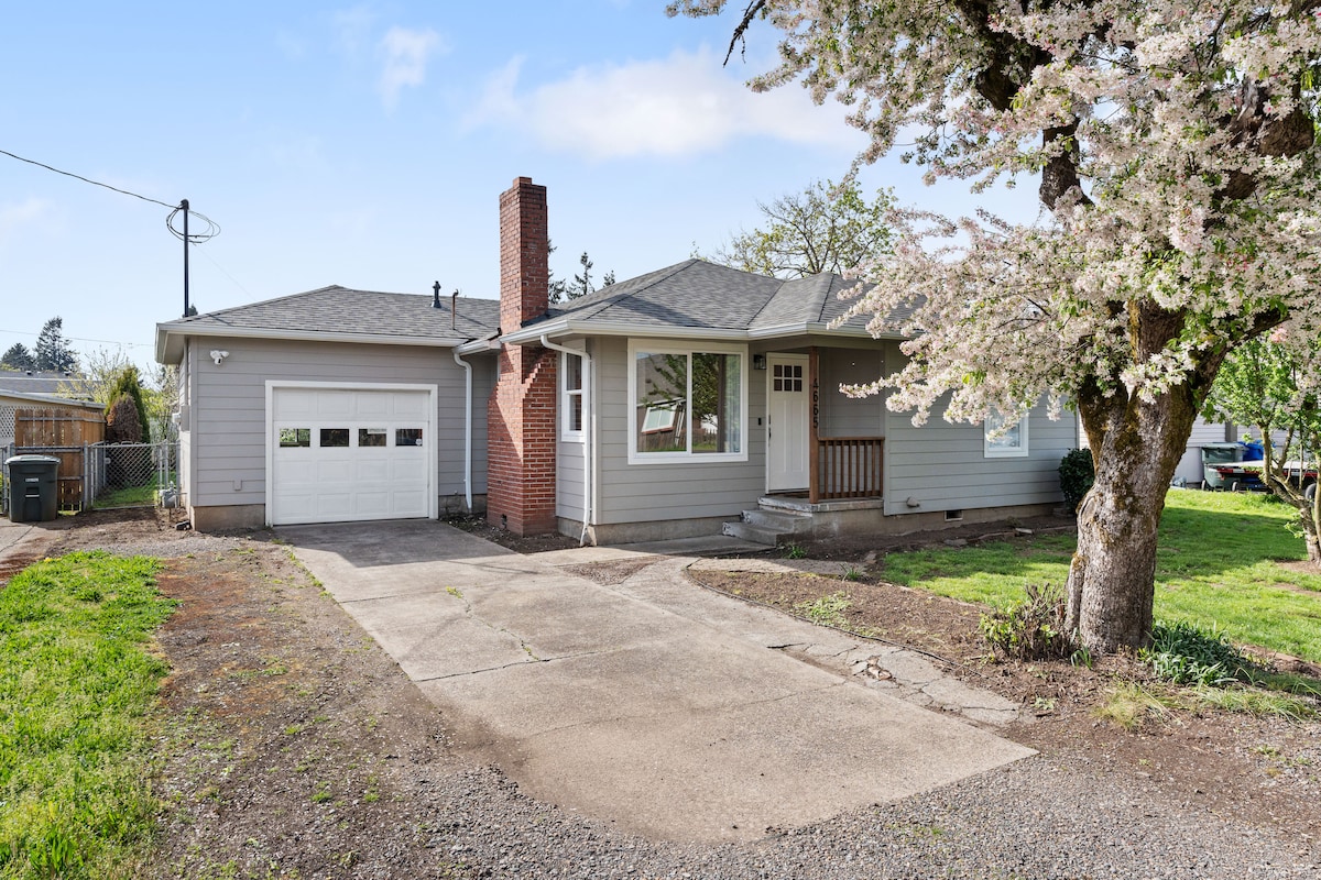 A single-story home is depicted with a light grey exterior and a chimney. A driveway leads to a garage, while a blooming tree adds a natural element to the front yard. The surrounding area features grassy spaces and pathways.
