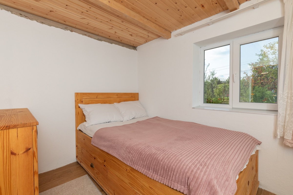A cozy bedroom features a wooden bed with a soft pink blanket and fresh white pillows. Natural light enters through a window, illuminating the room, which also includes a matching wooden dresser. The walls are painted white, and exposed wooden beams enhance the rustic charm.