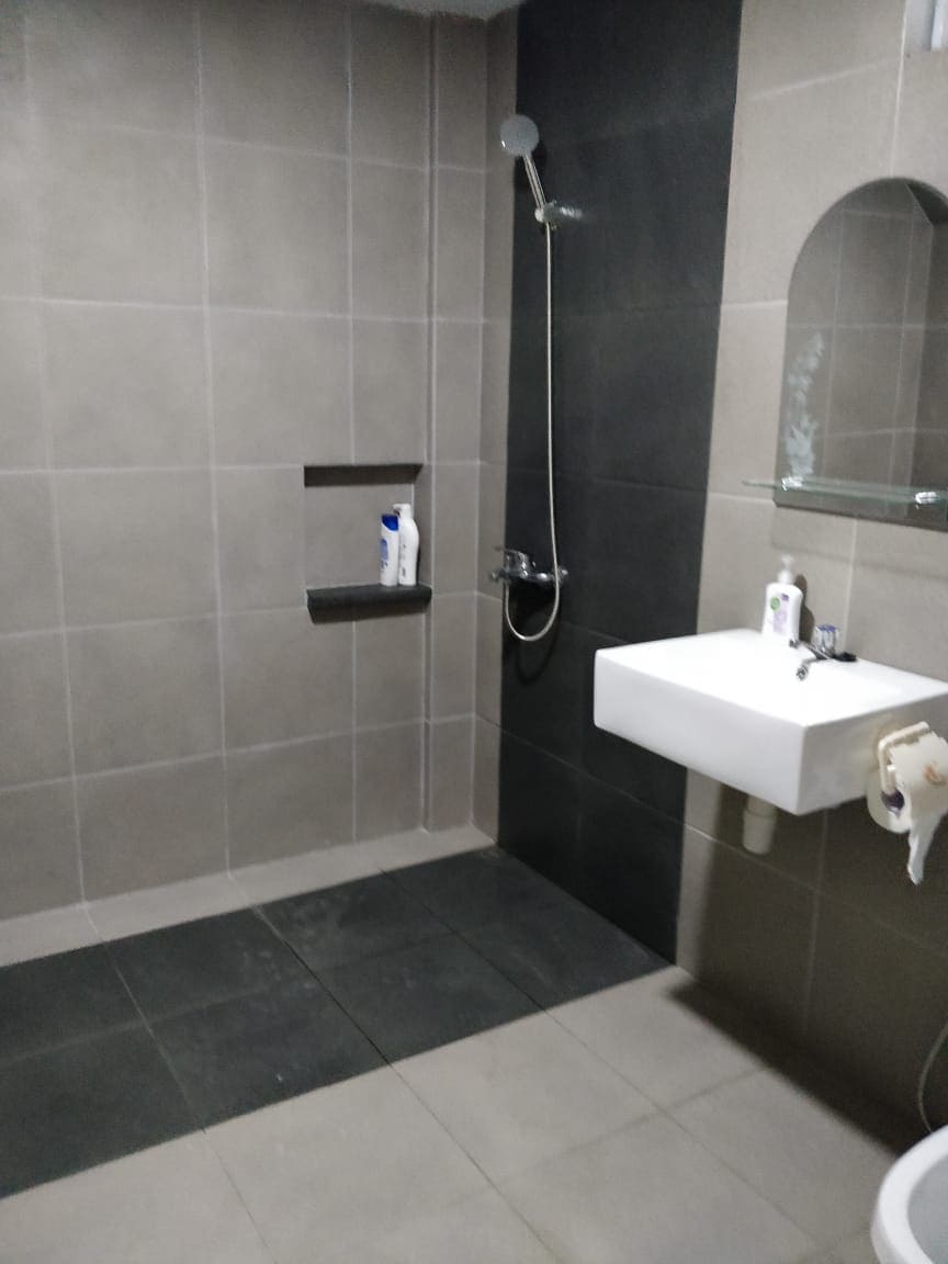 A modern bathroom is presented, featuring neutral-toned tile walls and flooring. A spacious shower with a rain showerhead is visible alongside a wall-mounted sink. Shelving built into the wall holds toiletries, and a small mirror is mounted above the sink.