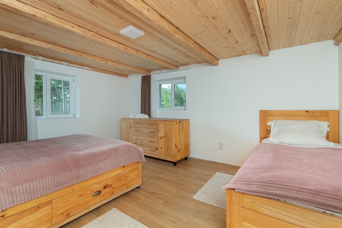 The spacious family bedroom features wooden beams and light wood paneling. A comfortable double bed and a single bed are arranged within the room. Natural light fills the space through two windows, with a simple wooden dresser positioned nearby.