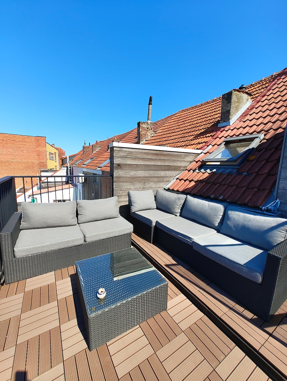 A spacious terrace is shown with modern outdoor seating arranged around a square table. The area features comfortable gray cushions and a warm wooden deck, providing an ideal spot for relaxation while enjoying clear blue skies.