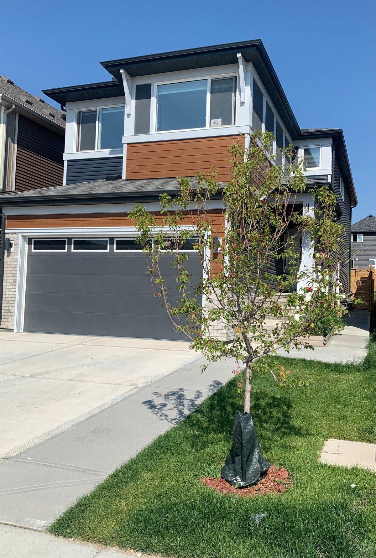 The exterior of a modern two-story home is depicted, featuring a combination of wood and stone cladding. A driveway leads to a garage door, while a small tree stands in the front yard, surrounded by green grass and showcasing a clear blue sky.