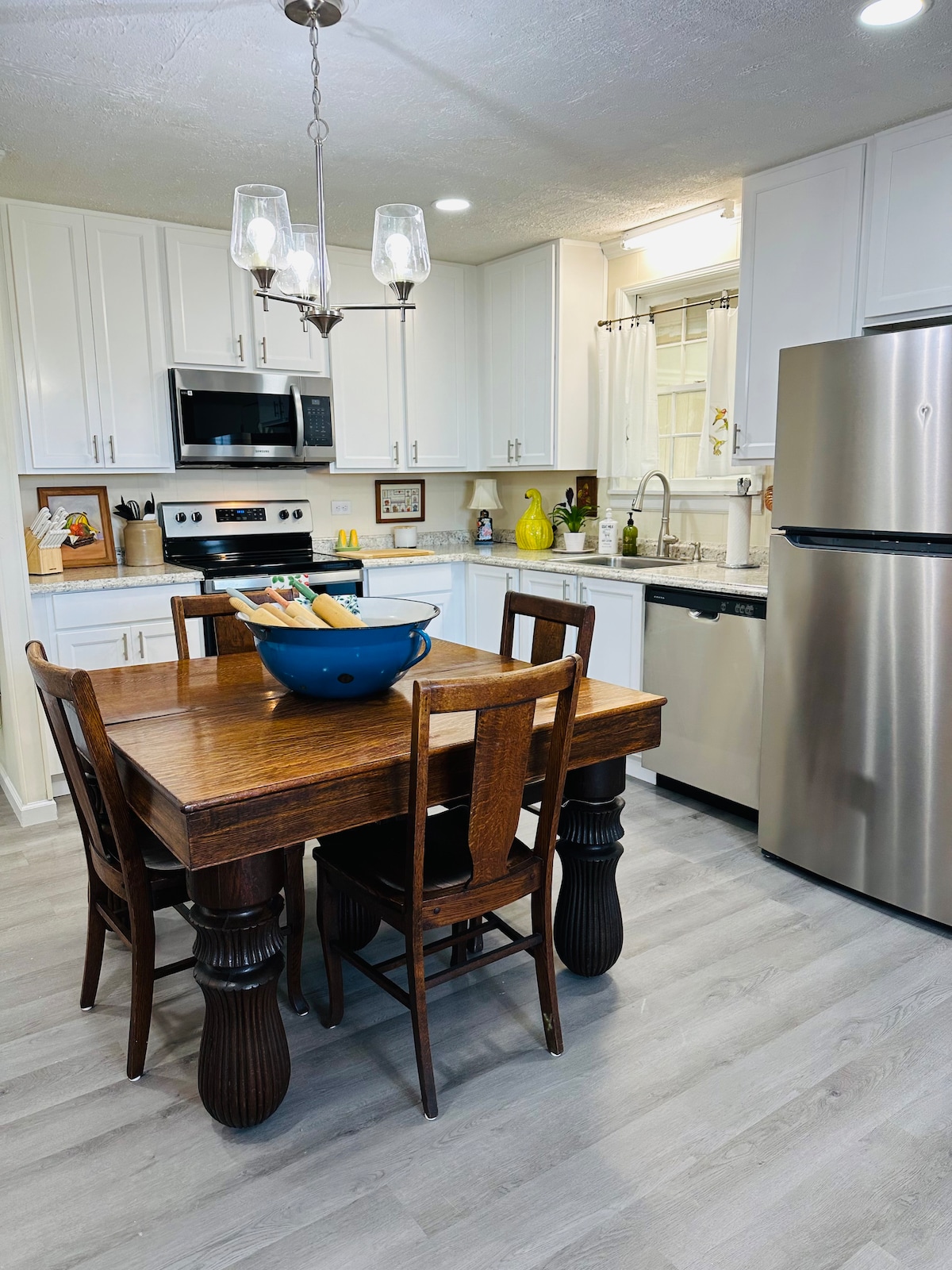 A kitchen area is depicted, featuring crisp white cabinetry and modern stainless steel appliances. A wooden table with four matching chairs sits in the center, complemented by a deep blue bowl atop it. Ample counter space and warm lighting create a functional, inviting environment.