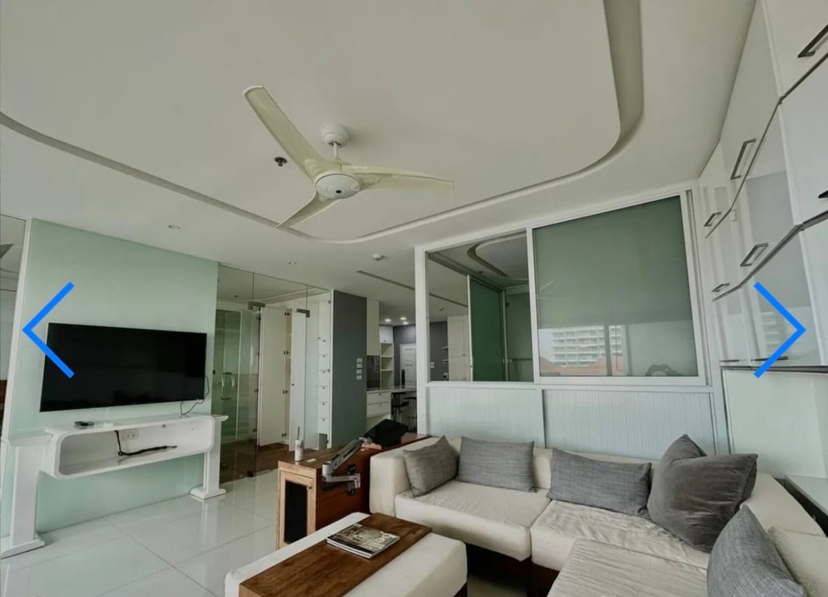 A modern living area features a spacious sectional sofa adorned with gray pillows. A ceiling fan is overhead, and a flat-screen television is mounted on the wall. Large glass windows provide a view of the surrounding area, creating a bright and airy atmosphere.